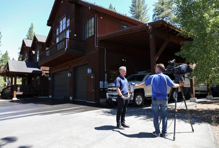 A man interviewing and video taping a man at a media event. They are standing in front of a home where a live demonstration of a digitally connected sprinkler system took place.