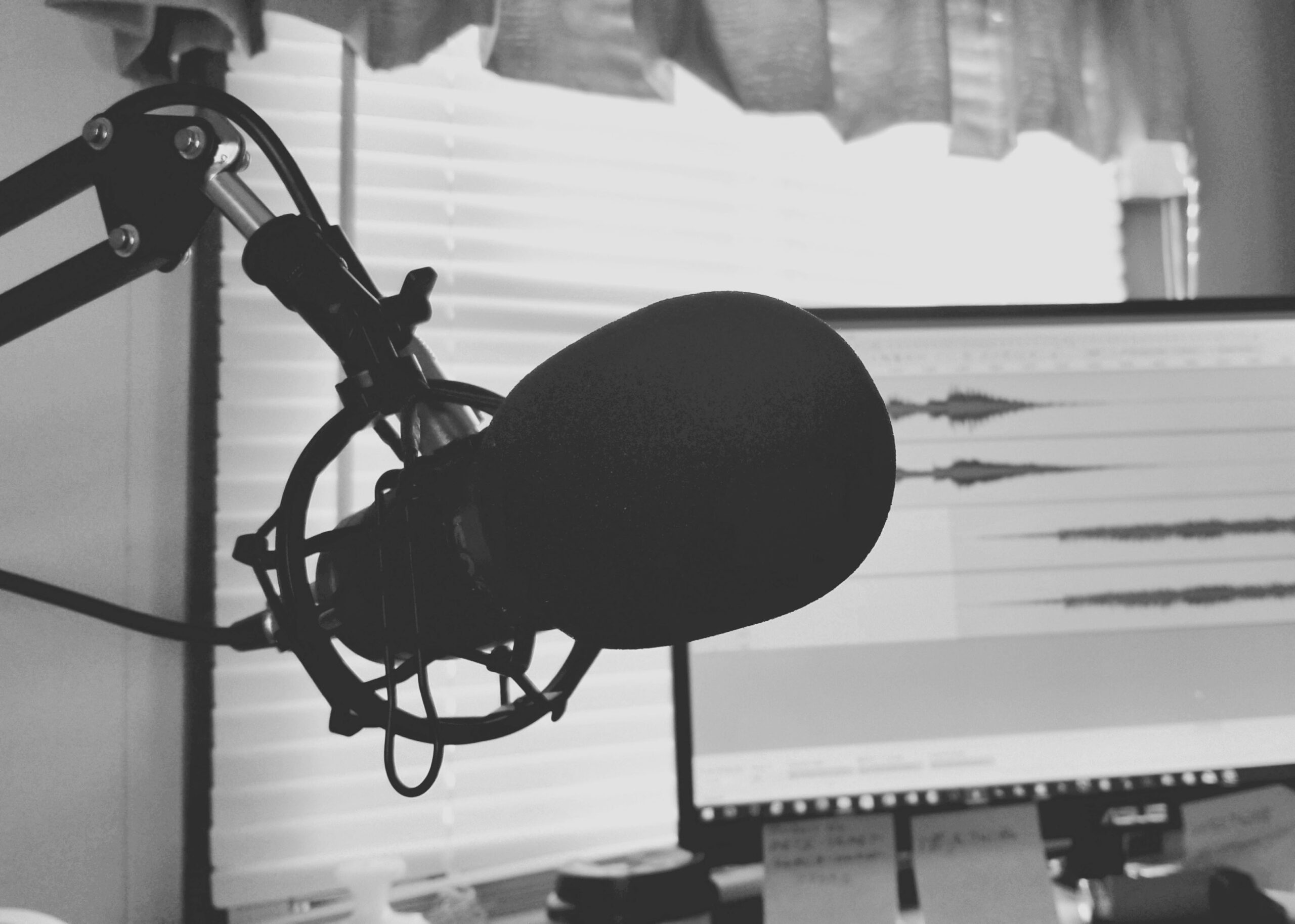 A podcasting set up with a microphone and a computer editing in the background.