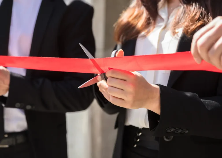 a woman's hands holdinc scissors and cutting a red grand opening ribbon while two men help hold the ribbon