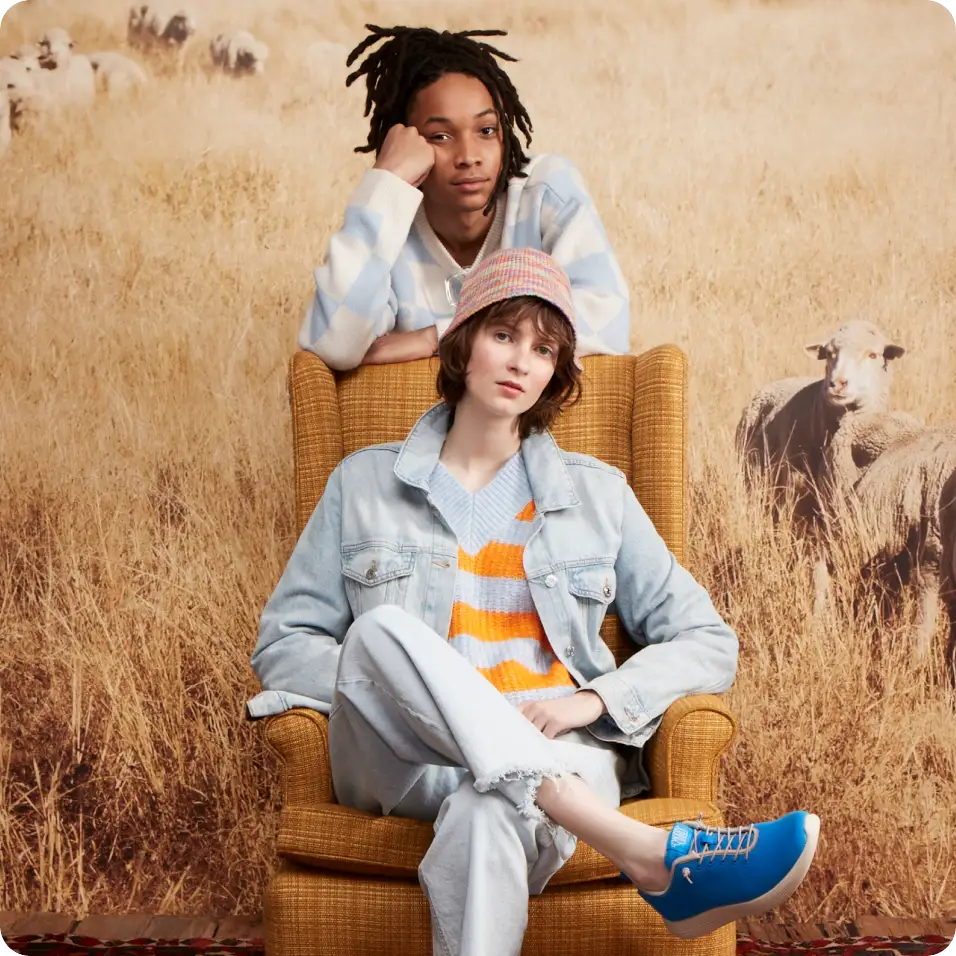 a woman in a chair wearing a blue sneaker and a person behind her in a field of grasses and sheep