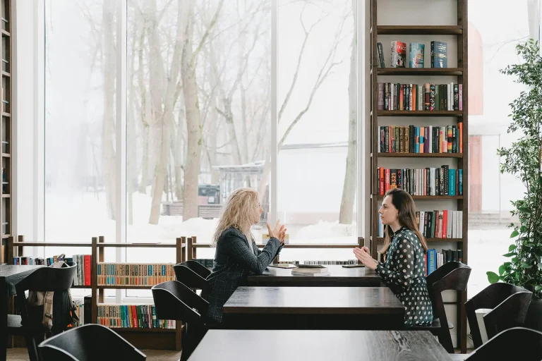 two women at a table in a library with snow outside the large window
